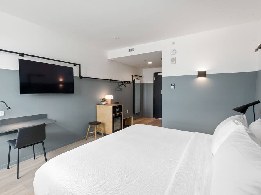 A modern hotel room with a bed, a desk, a TV, and a small kitchenette.