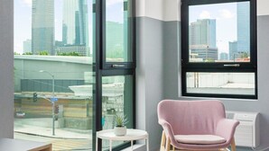 A modern room with large windows offering a city view, a pink chair, and a small table with a plant. - The Metric (Los Angeles)