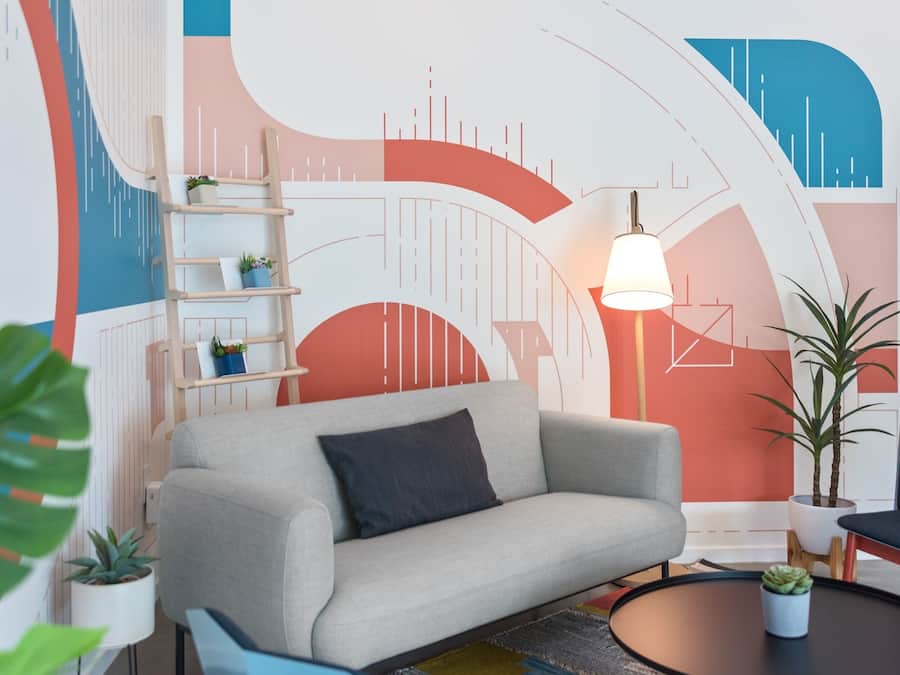 A modern living room with a grey sofa, a wooden ladder shelf with plants, a round coffee table, and a vibrant abstract wall mural.