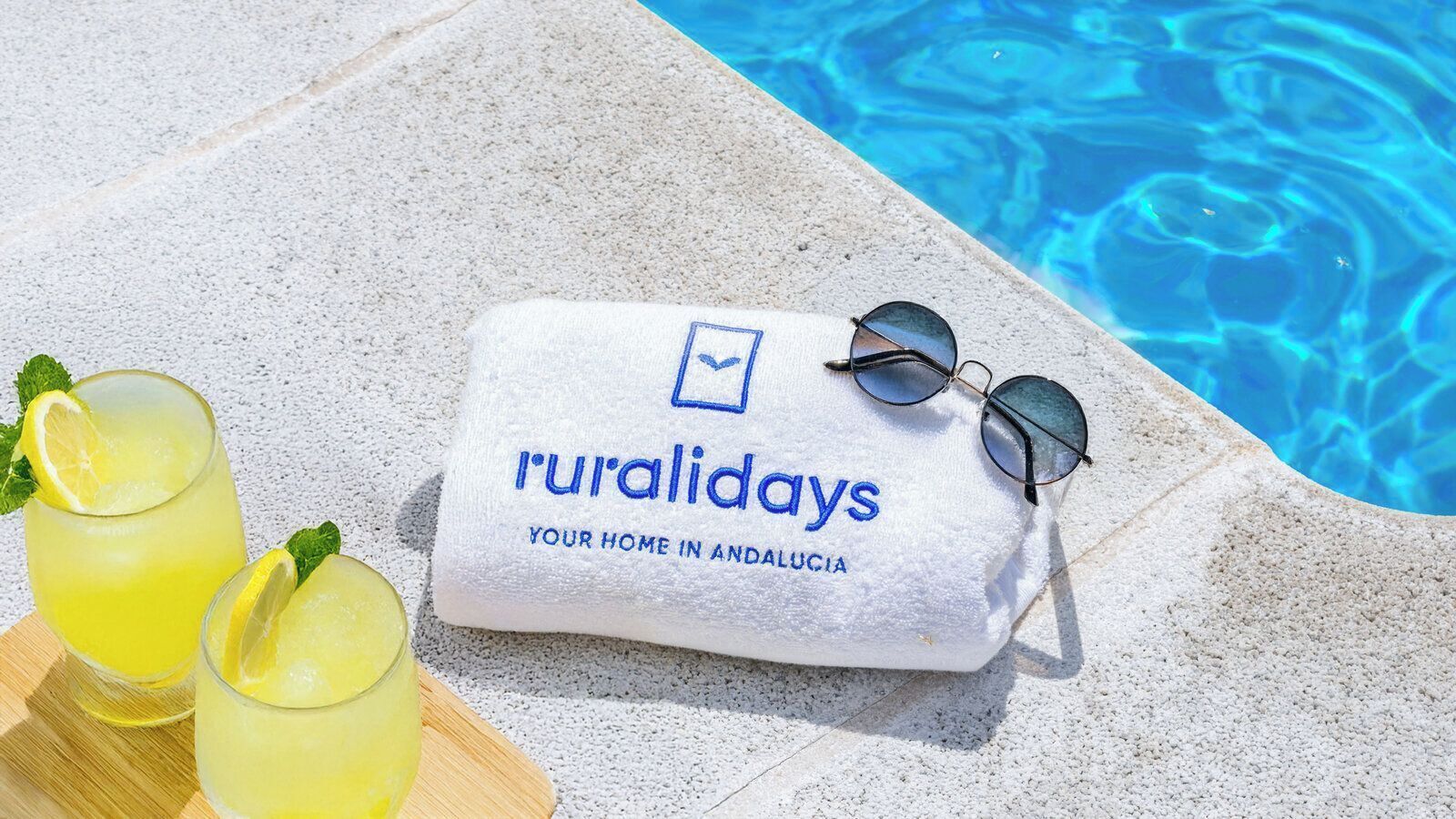 Una toalla con el texto 'ruralidays' y 'tu hogar en Andalucía', gafas de sol y dos vasos de una bebida amarilla con rodajas de limón junto a una piscina.