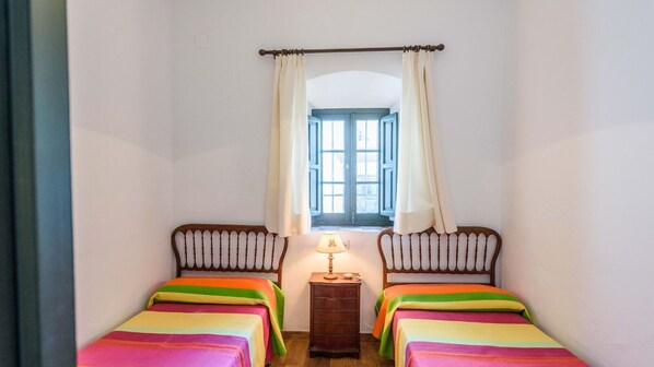Une petite pièce avec deux lits, une table de chevet, une fenêtre avec des rideaux et une lampe.