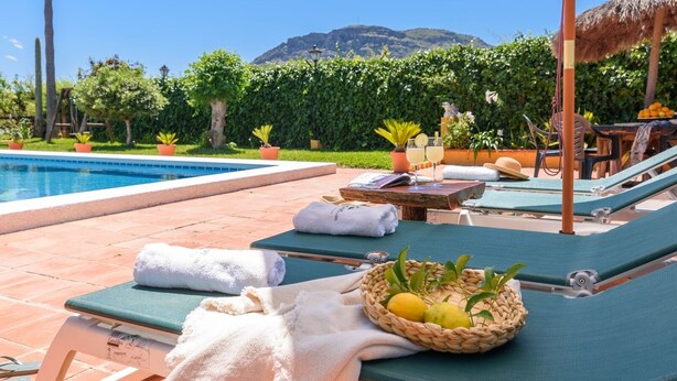 Zona de piscina amb gandules, tovalloles, una cistella de fruita i una taula amb una beguda i un llibre.