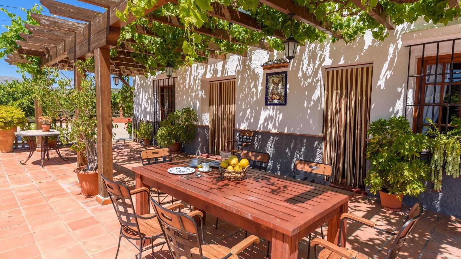An outdoor patio with a wooden table set for dining, surrounded by chairs, and potted plants.