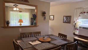Un comedor con una mesa de madera dispuesta para cuatro personas, rodeada de sillas. Hay un ventilador de techo encima de la mesa, y se ve una ventana con cortinas.