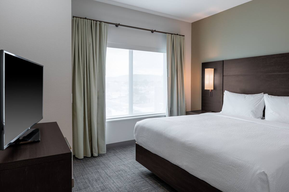 A hotel room with a large bed, a flat-screen TV on a wooden dresser, a window with curtains, and a nightstand with a lamp.