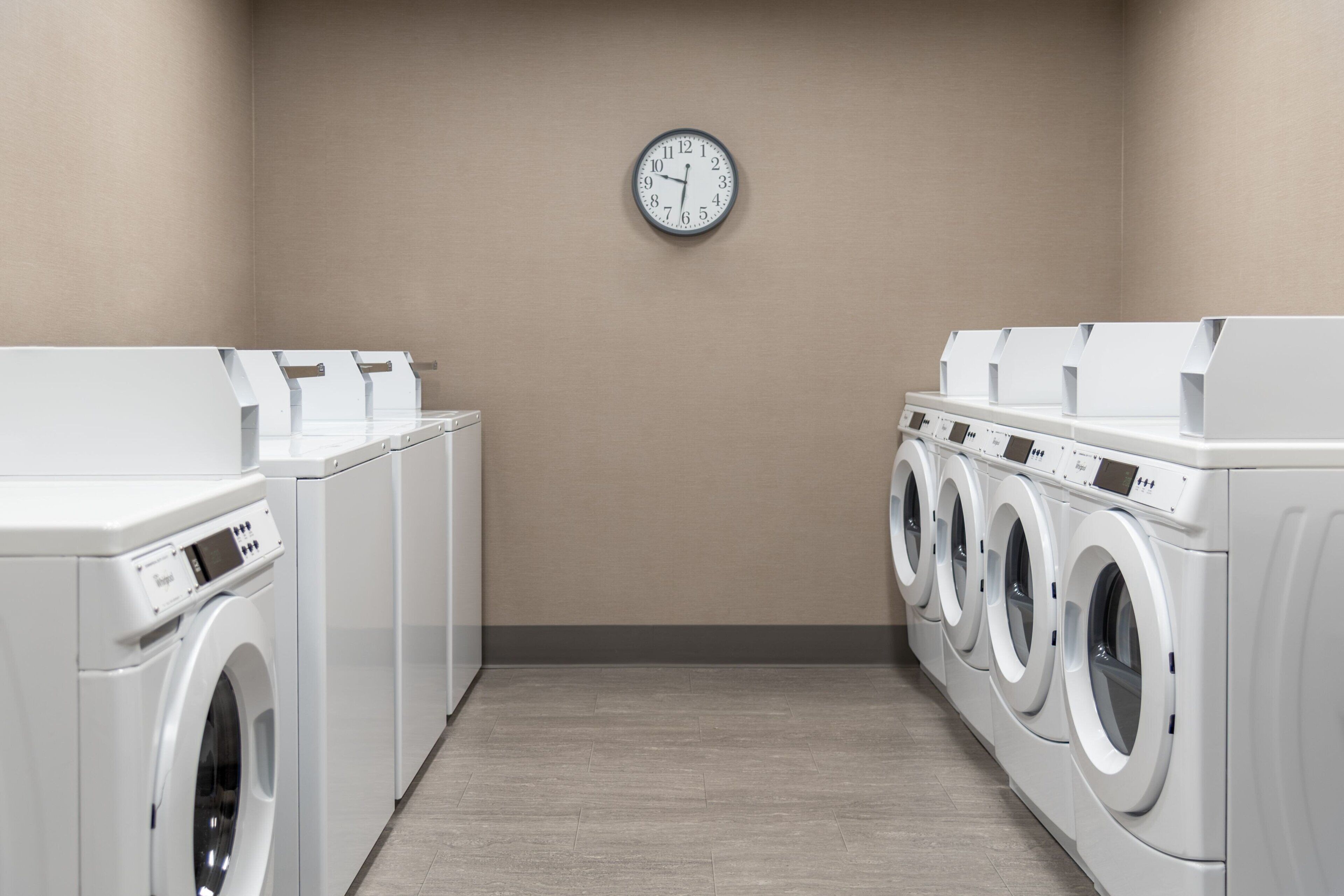 A laundry room with multiple washing machines, a clock on the wall, and a beige wall.