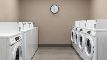 A laundry room with multiple washing machines, a clock on the wall, and a beige wall.