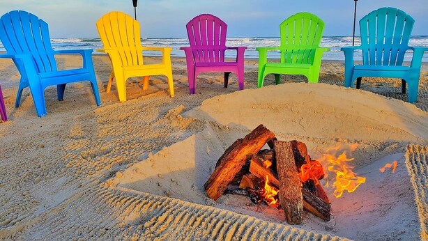 Eine Reihe bunter Adirondack-Stühle an einem sandigen Strand, inmitten davon eine Feuerstelle.