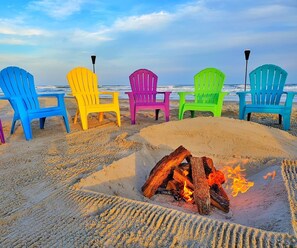 A row of colorful Adirondack chairs facing a beach with a fire pit in the sand.
