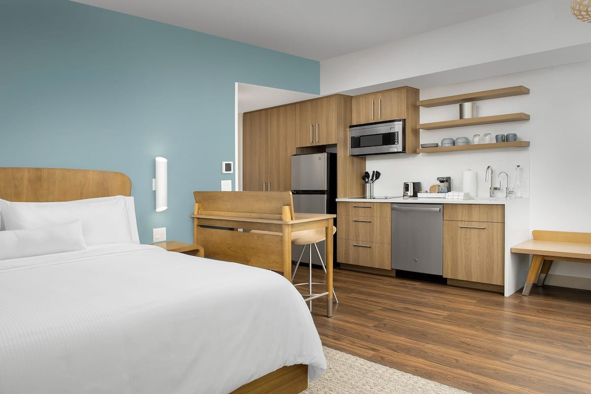 A modern hotel room with a bed, a desk, a kitchenette, and a bathroom.