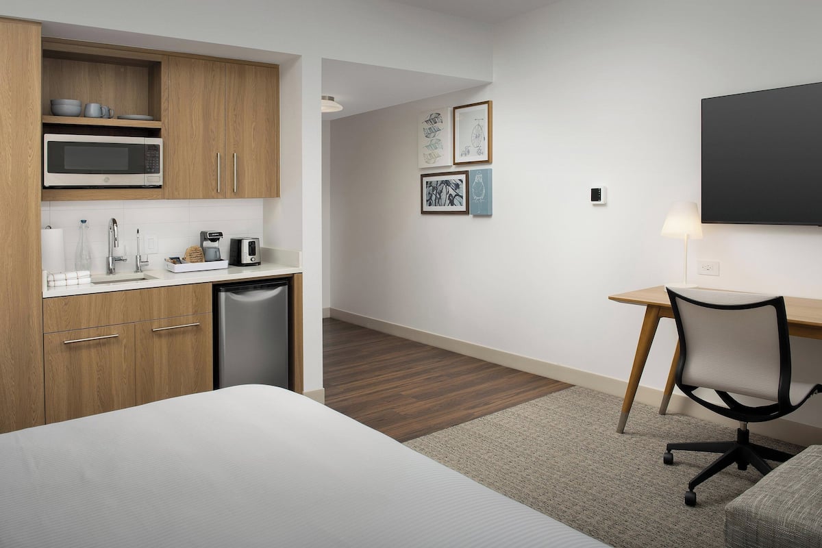 A modern hotel room with a kitchenette, a bed, a desk, and a chair.