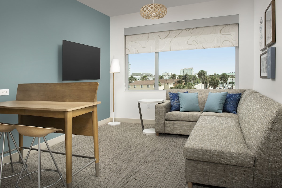 A modern living room with a wooden dining table, two bar stools, a grey sofa with blue cushions, a white floor lamp, and a large window with a view of buildings and palm trees.