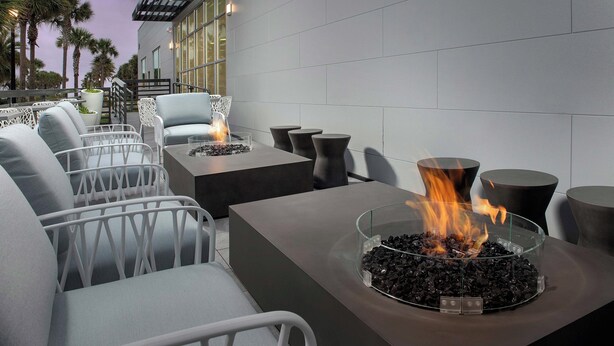 A modern outdoor seating area with white wicker chairs, a glass fire pit with burning wood, and a view of palm trees and a building in the background. - Element by Marriott Jacksonville Beach (Jacksonville Beach)