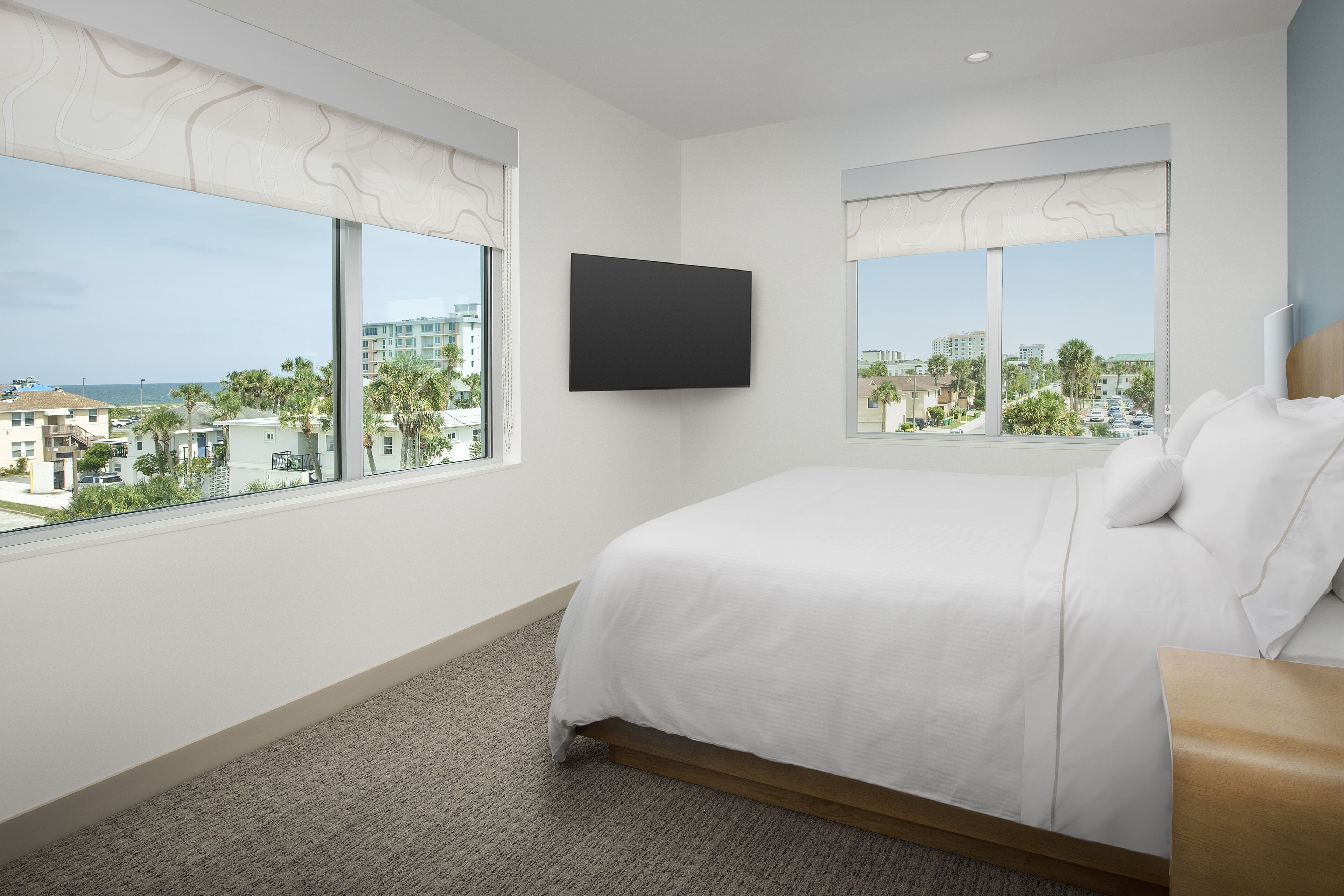 A hotel room with a large bed, two windows with curtains, a flat-screen TV, and a view of the city and beach.