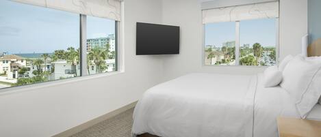 A hotel room with a large bed, two windows with curtains, a flat-screen TV, and a view of the city and beach.