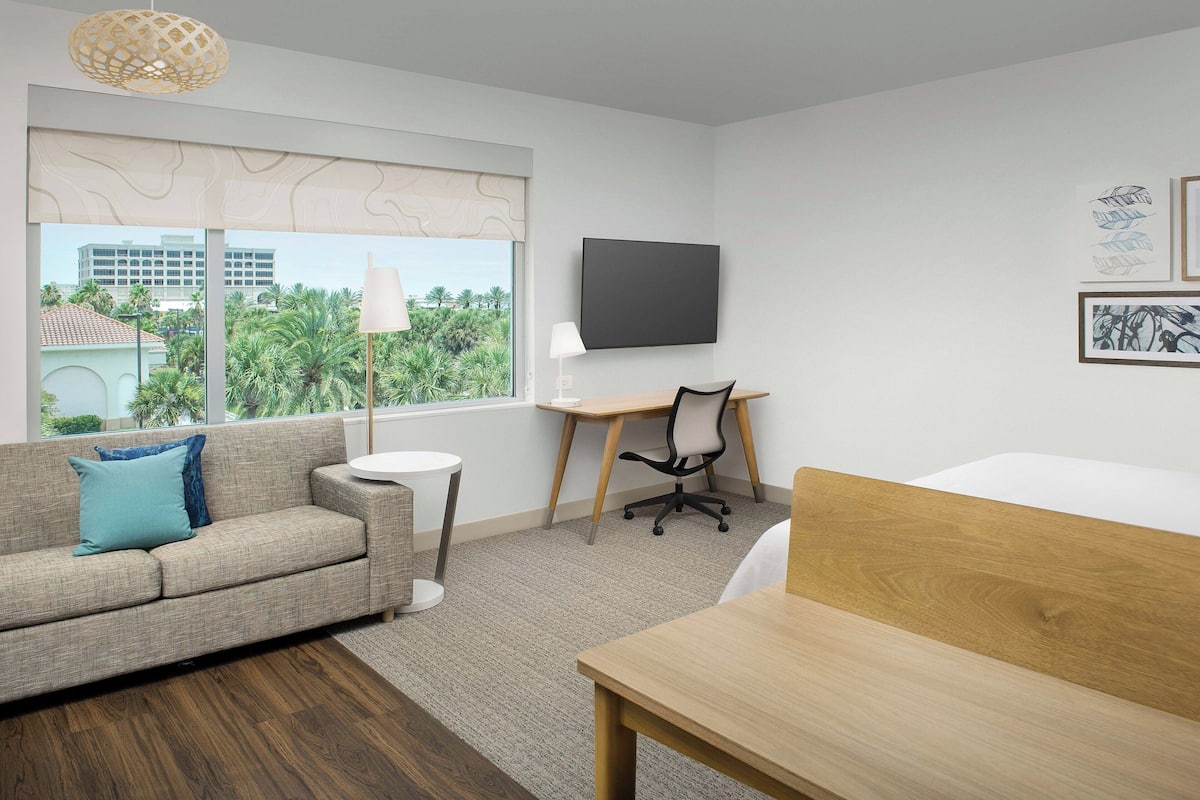 A modern hotel room with a sofa, a desk, a bed, a TV, and a window with a view of palm trees and buildings.