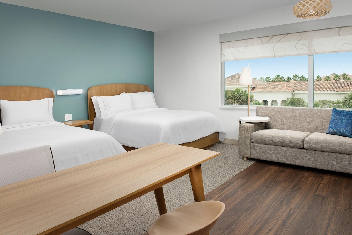 A hotel room with two beds, a sofa, a small table, and a view of buildings through a window.