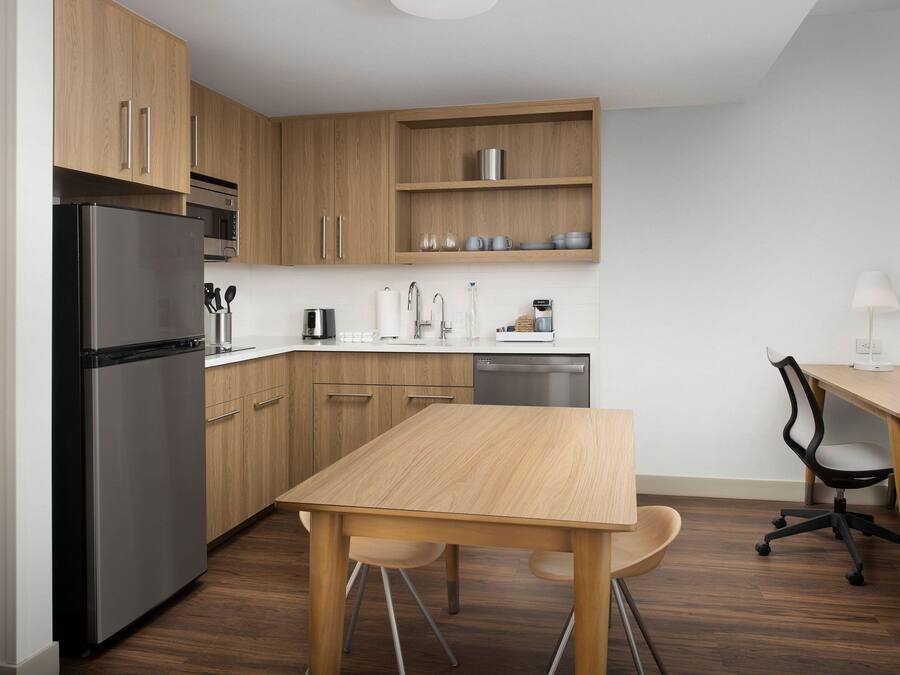 Ett modernt kök med träskåp, ett kylskåp i rostfritt stål, en mikrovågsugn och en diskmaskin. Här finns också ett matbord i trä med stolar och en liten kontorsstol.