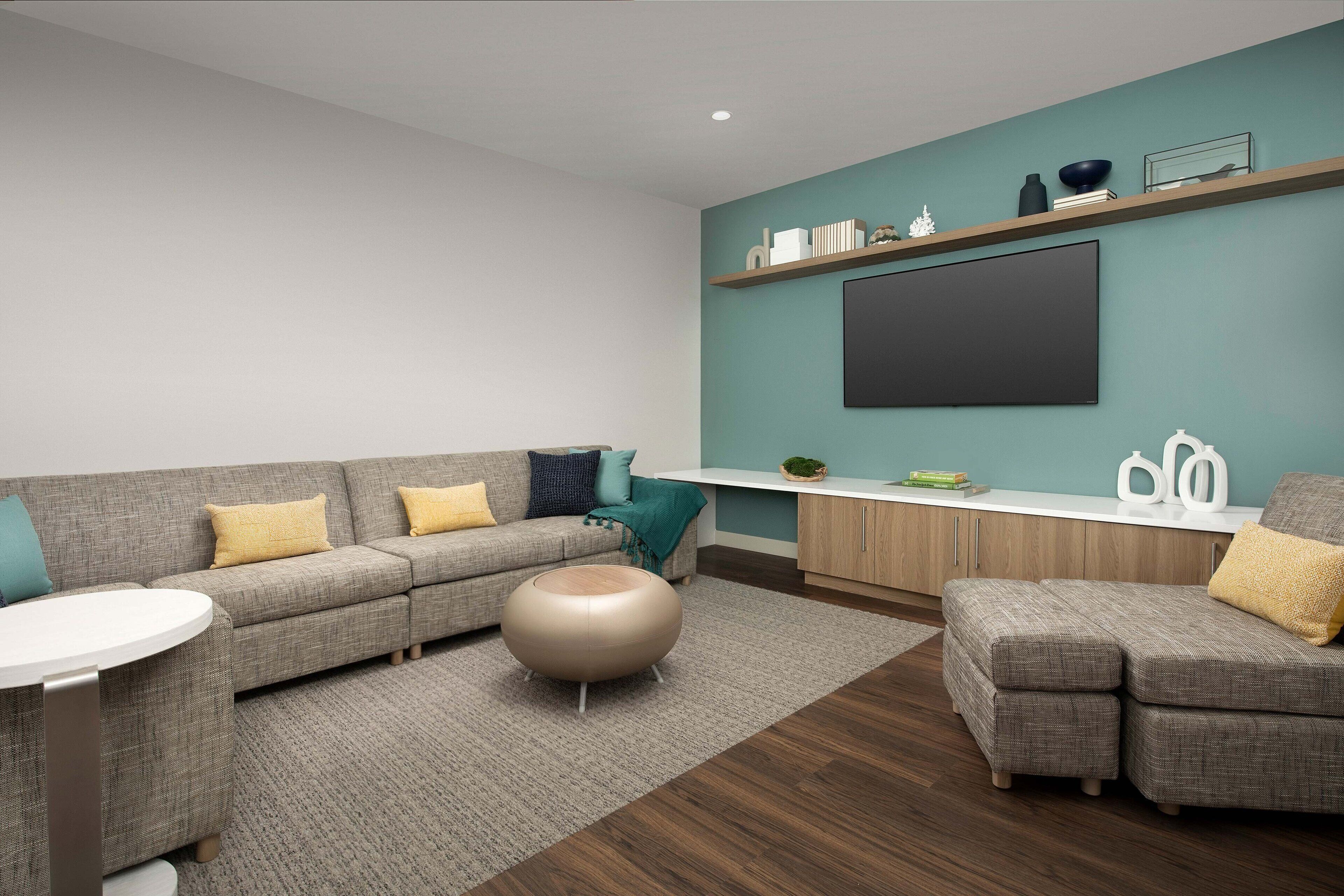 A modern living room with a grey sofa, a round ottoman, a small white table, and a teal accent wall.