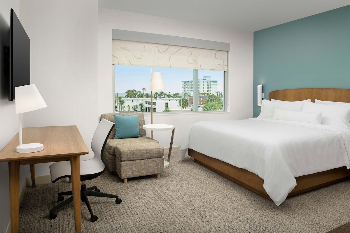 A hotel room with a bed, a desk, a chair, a small table, and a window with a view of buildings and palm trees.