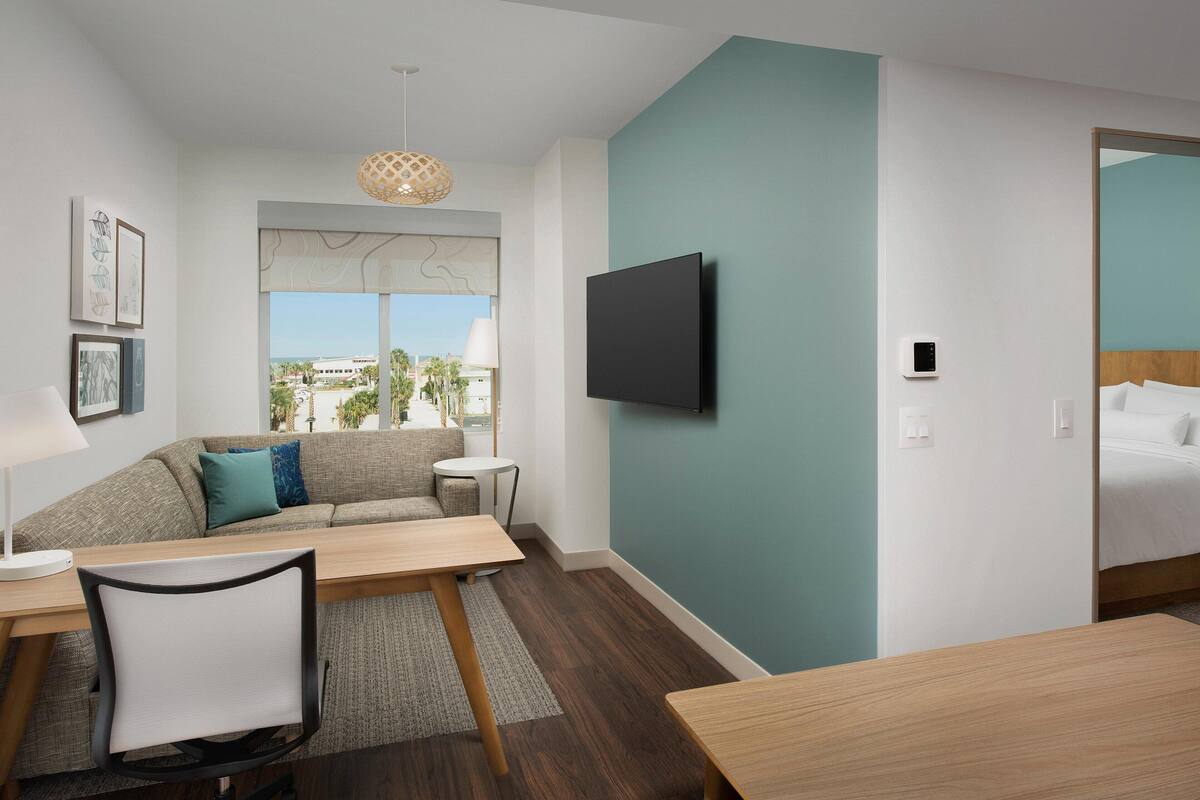 A modern hotel room with a sofa, a dining table, a TV mounted on the wall, and a view of the outdoors.