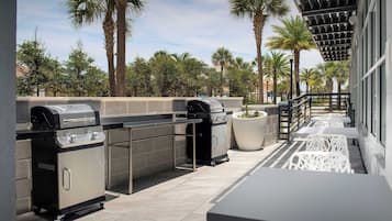 An outdoor patio with two barbecue grills, a seating area with white chairs, and a view of palm trees and buildings in the background.