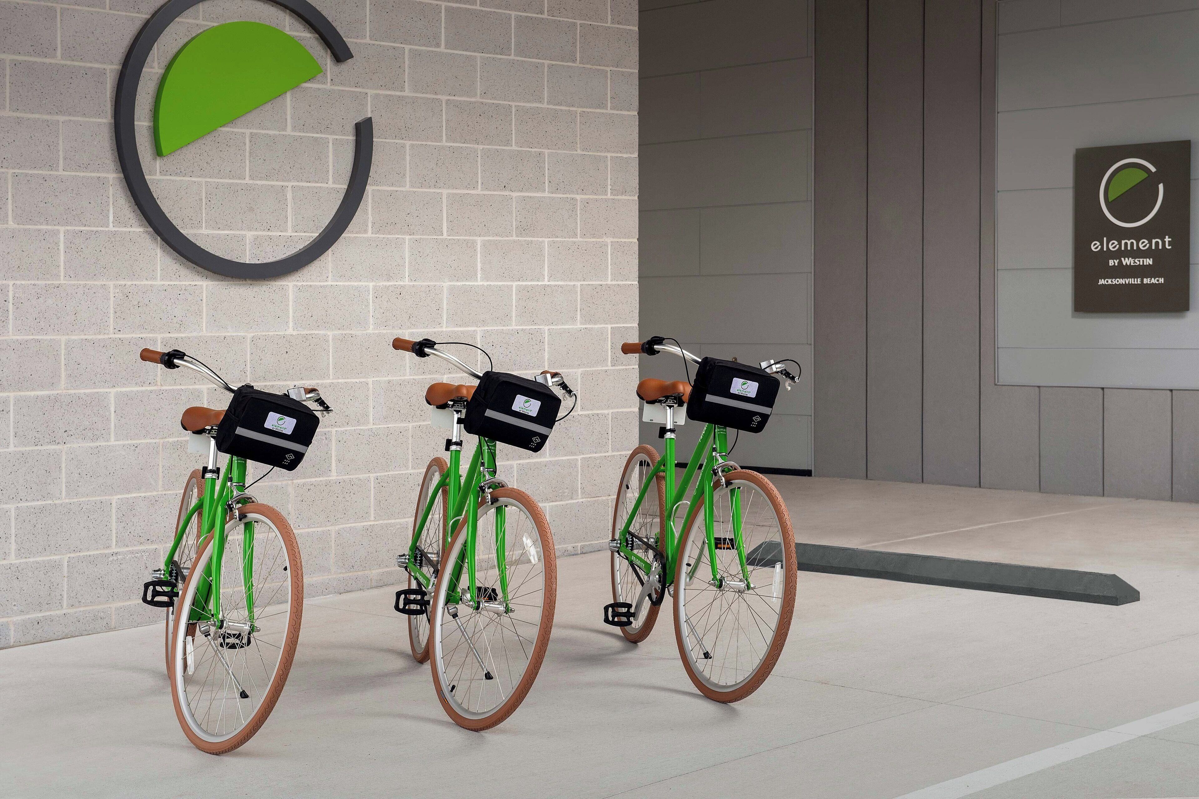 Three green bicycles with brown seats and black bags, parked against a wall with a large circular logo and a smaller sign that reads "element by Westin Jacksonville Beach".