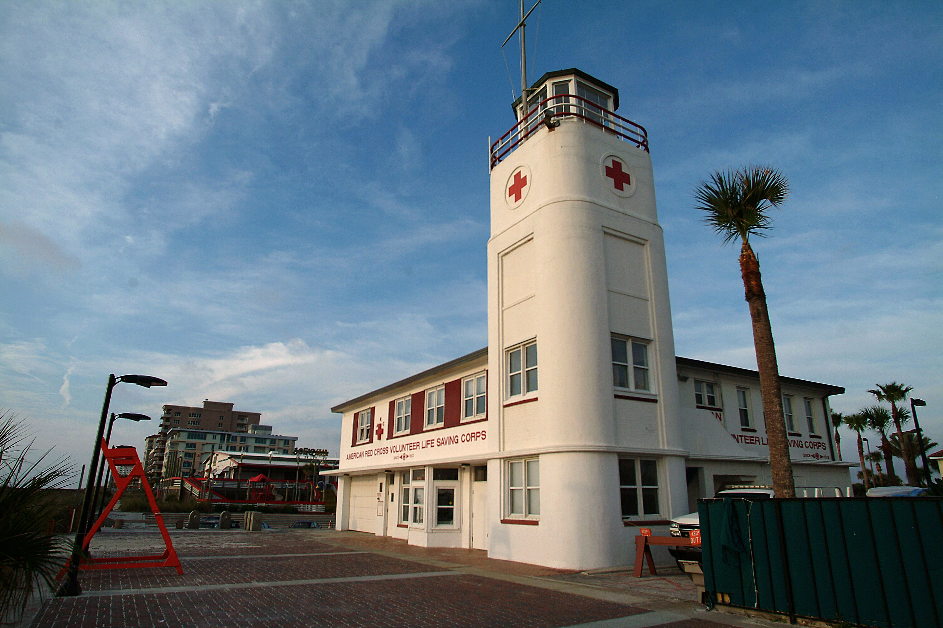 A building with a red cross symbol, indicating it is a medical facility, with a clear sky and palm trees in the background.