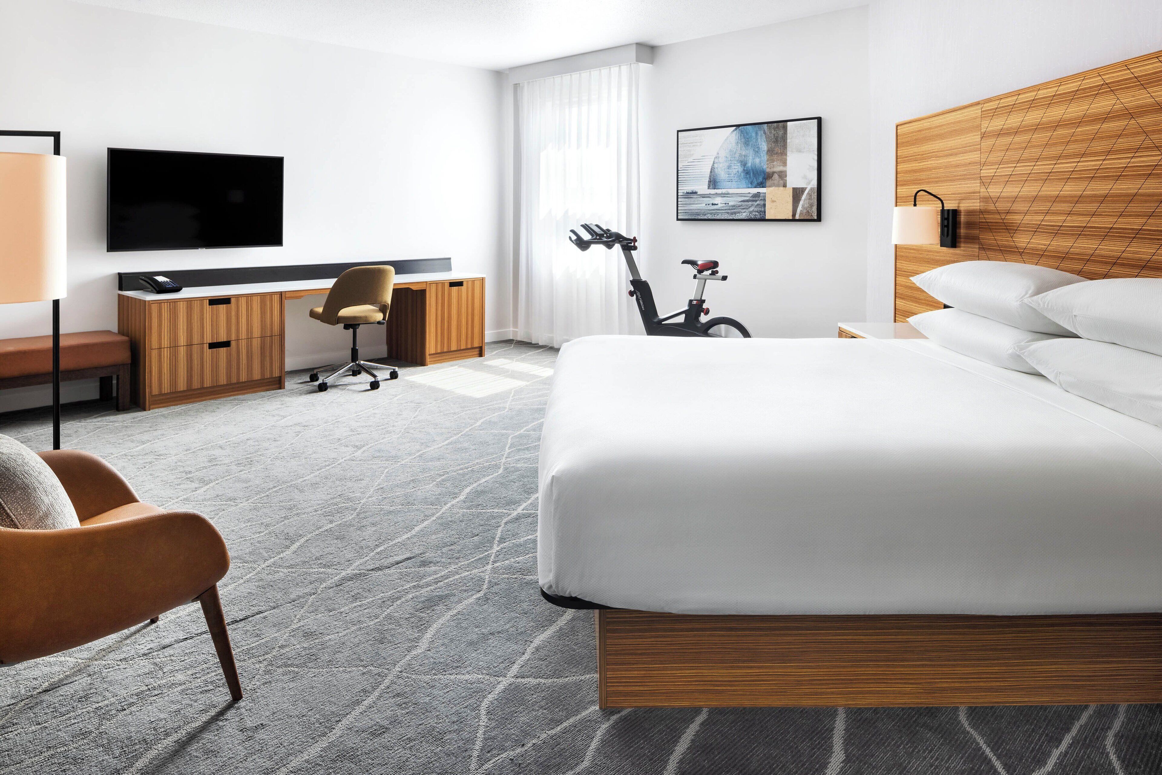A modern hotel room with a large bed, a desk with a chair, a television, and a bicycle.
