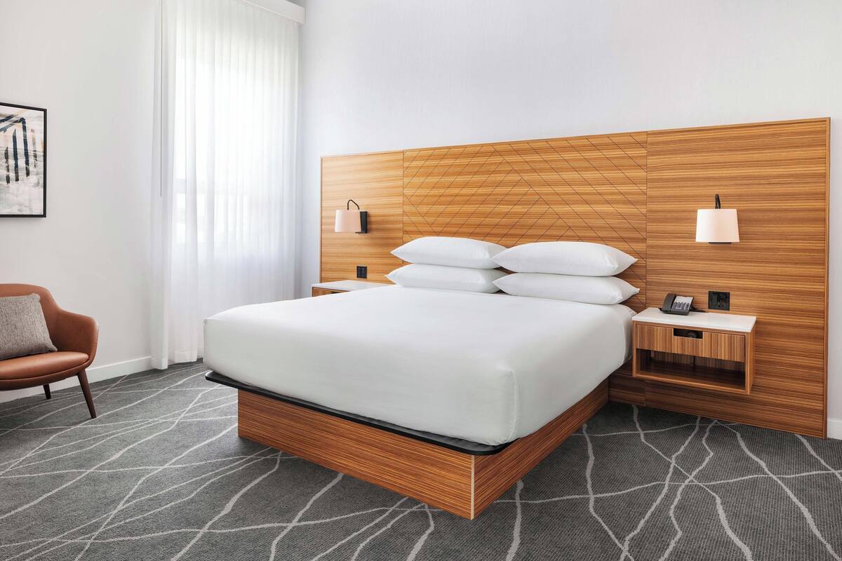 Une chambre d’hôtel moderne avec un grand lit, une tête de lit en bois, des tables de chevet avec des lampes, une chaise et une moquette à motifs.