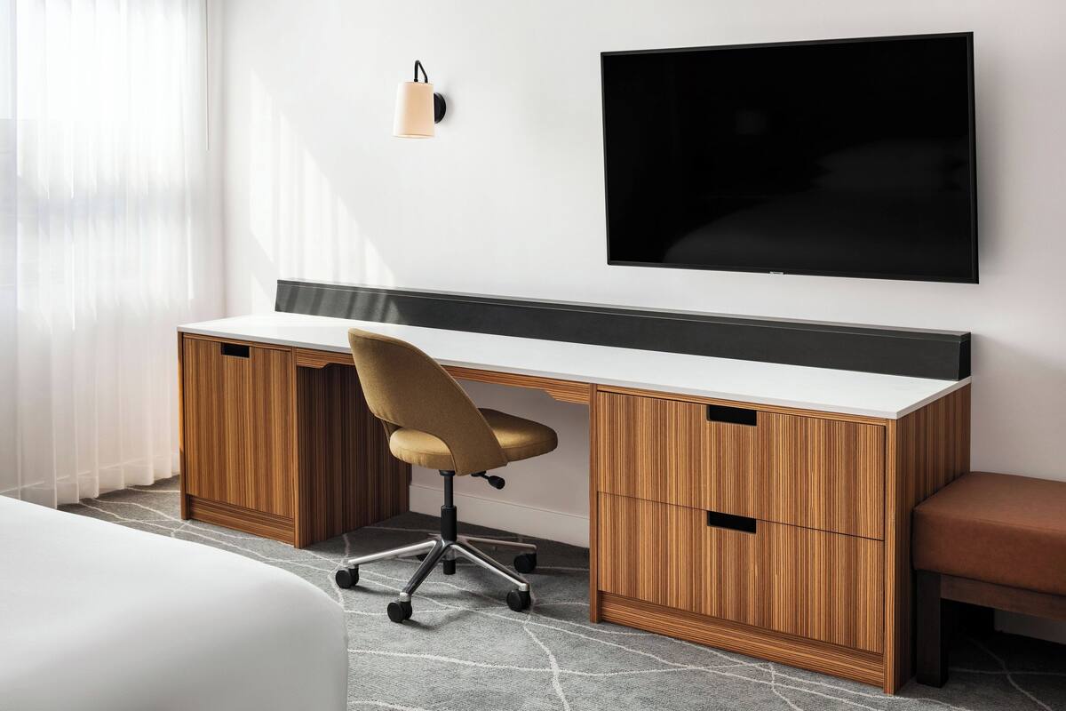 Une chambre d’hôtel moderne avec un bureau en bois, un fauteuil confortable, un téléviseur à écran plat fixé au mur et une petite table de chevet.