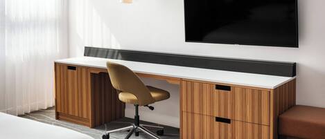 A modern hotel room with a wooden desk, a comfortable chair, a flat-screen TV mounted on the wall, and a small bedside table.