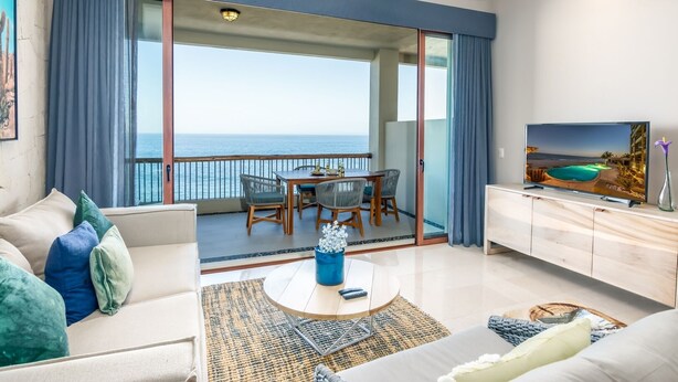 Ein modernes Wohnzimmer mit Balkon und Meerblick, einem Essbereich und einem Fernseher auf einem Holzschrank.
