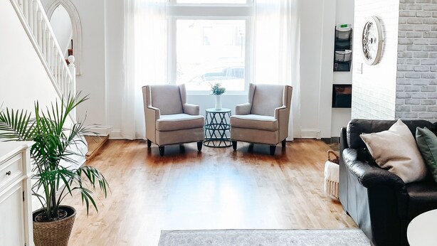 Ein geräumiges Wohnzimmer mit hoher Decke, einer Treppe links, einem schwarzen Ledersofa, einem gemusterten Teppich und einer Topfpflanze.