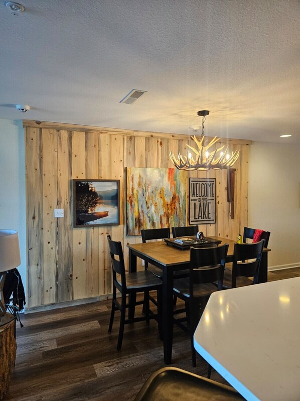 A dining area with a wooden wall, a painting, a 'Welcome to the Lake' sign, and a chandelier.