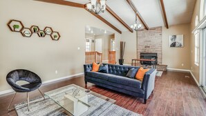 A spacious living room with a dark blue sofa, a glass coffee table, and a black chair. There is a brick fireplace and a wooden staircase in the background. - POOL. Spacious 4BR/2B. SF Bay Area (Newark)