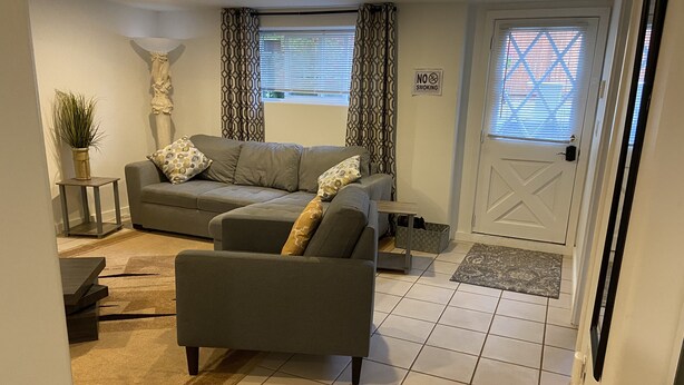 A living room with a grey sofa, a matching armchair, patterned curtains, a 'No Smoking' sign, and a door with a window. - Cozy 1 bedroom apartment, near Seattle & Airport. (Normandy Park)