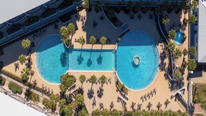 Oversigtsbillede af et resort med to swimmingpools, palmer og et overdækket område med siddepladser.