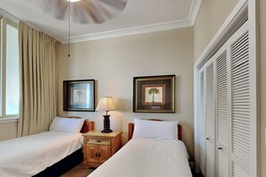 Habitación de hotel con dos camas individuales, una mesita de noche con lámpara, un ventilador de techo y un amplio ventanal con cortinas.
