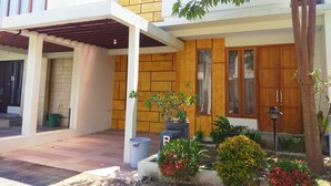 A two-story house with a covered porch, a wooden door, and a small garden with potted plants. - Omah Dixy Family Homestay by FH Stay (Baturetno)