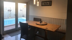 Uma área de refeições com mesa de madeira, cadeiras pretas e um banco. Uma piscina é visível pela porta de vidro.