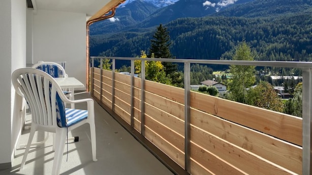 A balcony with a wooden railing, a white chair with blue stripes, and a view of snow-capped mountains and forested valleys. - Ferienwohnung STEINMANN by Interhome (Scuol)