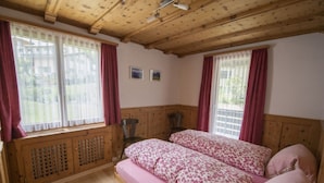 A bedroom with wooden paneling, two beds with floral bedding, a radiator, and a window with curtains. - Ferienwohnung STEINMANN by Interhome (Scuol)