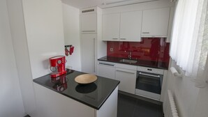 A compact kitchen with white cabinets, a black countertop, a red coffee maker, and a built-in oven. - Ferienwohnung STEINMANN by Interhome (Scuol)