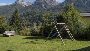 A grassy area with a wooden swing set and a ladder, surrounded by trees and mountains in the background. - Ferienwohnung STEINMANN by Interhome (Scuol)