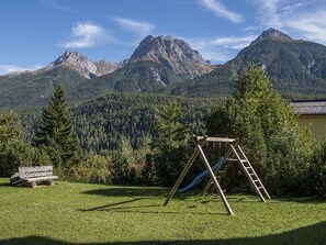 Relax on a well-maintained lawn with a swing set and slide, surrounded by a majestic mountain range and dense forest.