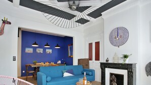 Een woonkamer met een blauwe bank, een roze fauteuil, een open haard, en een eetgedeelte met een tafel en stoelen.