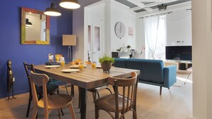 Een eetgedeelte met een houten tafel, stoelen en hanglampen. Achterin staat een blauwe bank en een televisie.