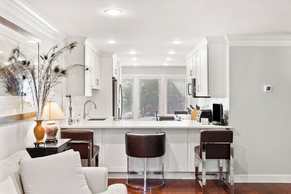 A modern kitchen with white cabinets, a central island with stools, a dining area with a white sofa, and a large window with blinds. - Modern and Spacious home with a fantastic, central, downtown location (Charleston)