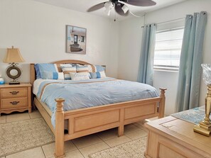 A bedroom with a wooden bed, a nightstand with a lamp, a painting on the wall, and a window with curtains. - Oceanview Across From The Beach (Ormond by the sea)
